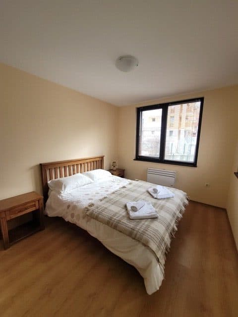 A simple bedroom with a double bed, two folded towels, wooden headboard, two bedside tables, and a window letting in natural light.