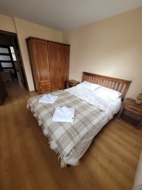 A neatly made double bed with a plaid blanket and two folded towels, wooden bedside tables, a wardrobe, and a wooden floor in a beige-walled bedroom.