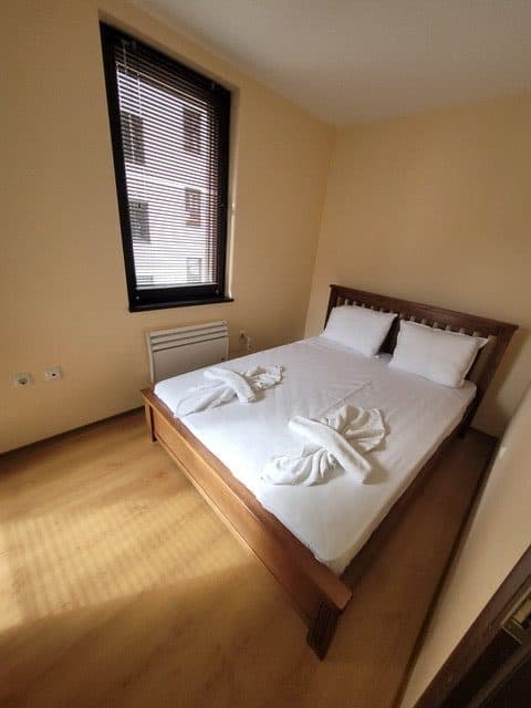 A small bedroom with a wooden double bed, white bedding, two pillows, two towels shaped like swans, a window with blinds, and light-colored walls and flooring.