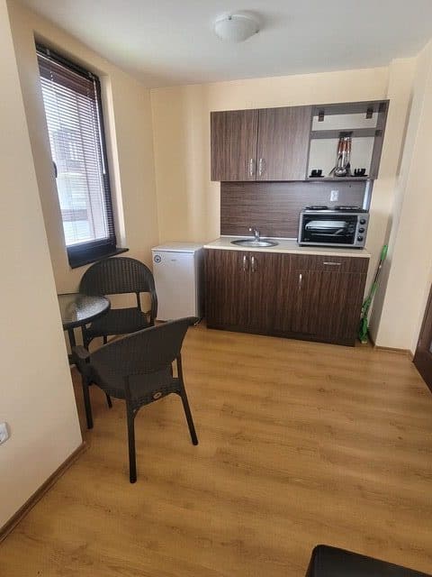 Compact kitchen with wood cabinets, a small refrigerator, microwave, sink, round table with two chairs, and a broom in the corner on a light wood floor.