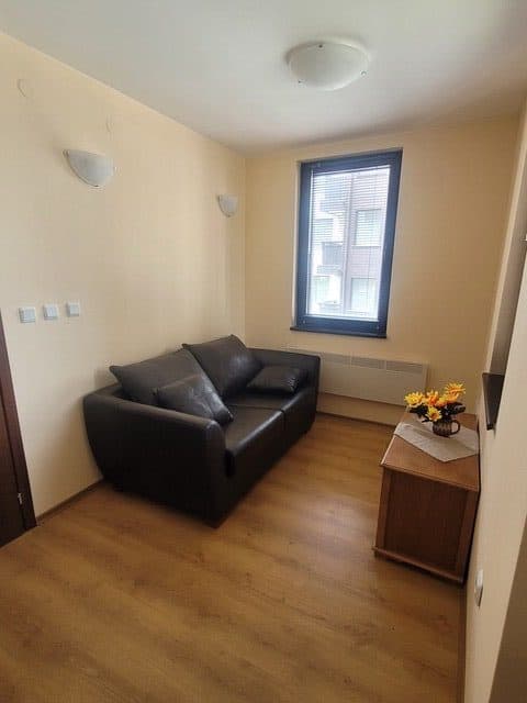 A small room with a black leather sofa, a window, a wall-mounted TV, and a wooden cabinet with a vase of yellow and orange flowers.