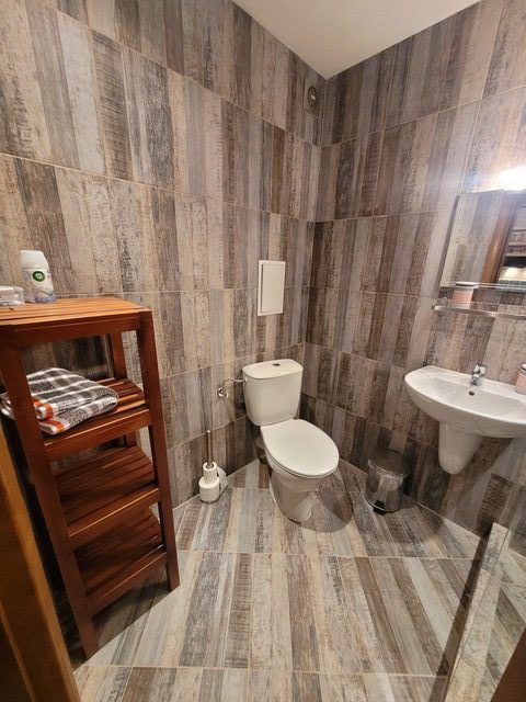 Bathroom with tiled walls and floor, featuring a toilet, sink, mirror, towel rack with folded towels, and toiletries on a wooden shelf.