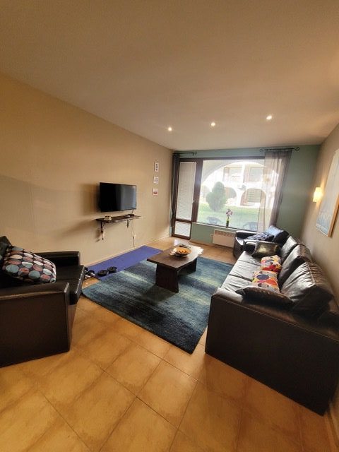 A living room with two black sofas, a wall-mounted TV, a coffee table, and a blue rug on a tiled floor. Large window and glass door let in natural light.