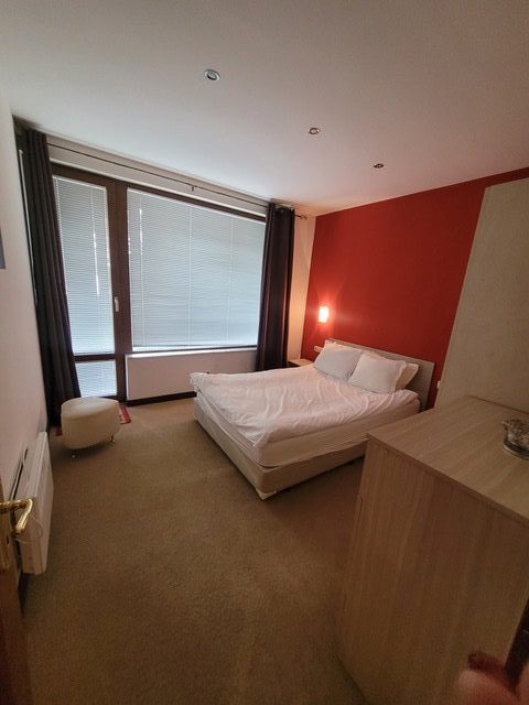 A small bedroom with a single bed, a nightstand, a dresser, a window with blinds, and beige carpet flooring.