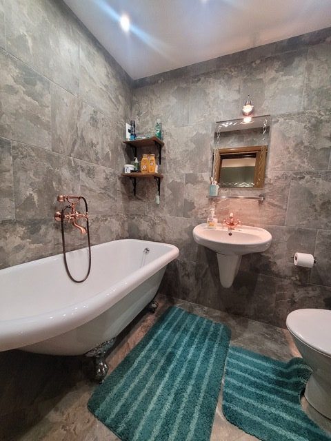 A bathroom with gray tile walls and floor, a freestanding bathtub, pedestal sink, mirror, toilet, teal striped rugs, and a small shelf with toiletries.