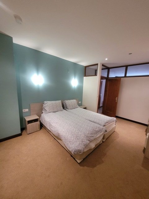 A simple bedroom with a double bed, two bedside tables, pale blue accent wall, beige carpet, and a wooden door at the far end of the room.